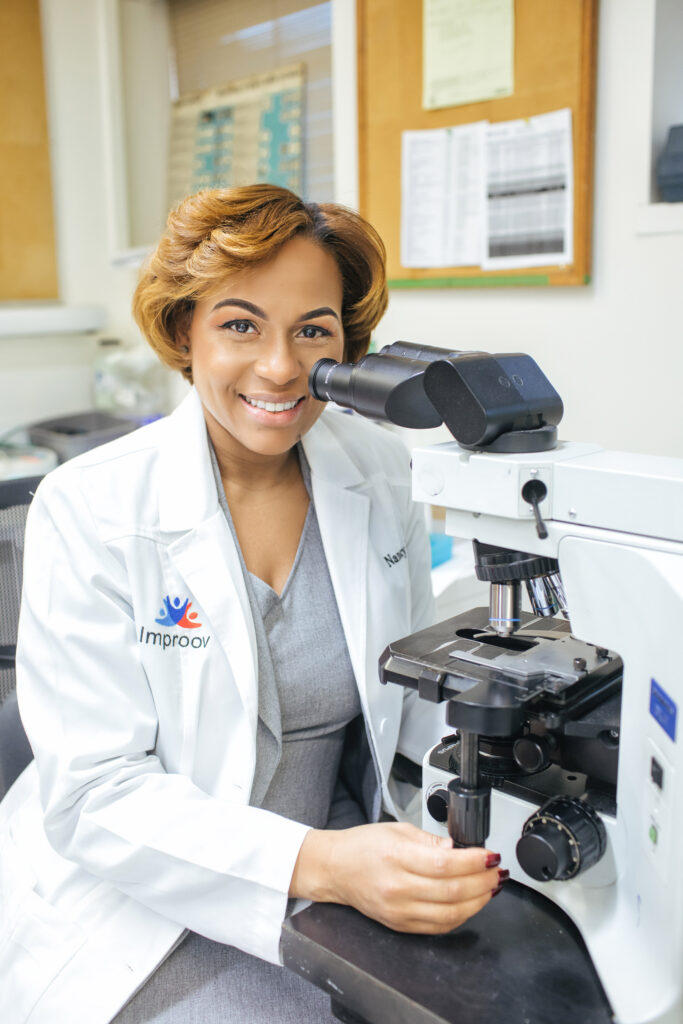 A Light-Skinned Black Woman wearing a white laboratory coat with the logo Improov Labs on the front of the coat. She is leaning over a microscope and smiling at the camera. Nancy Alers, MS, MT, (ASCP)cm, CQIA, CQ/OE, CLC, is the founder of Improov Labs, and the Assistant Director for Lab Operations and Quality at Montefiore.