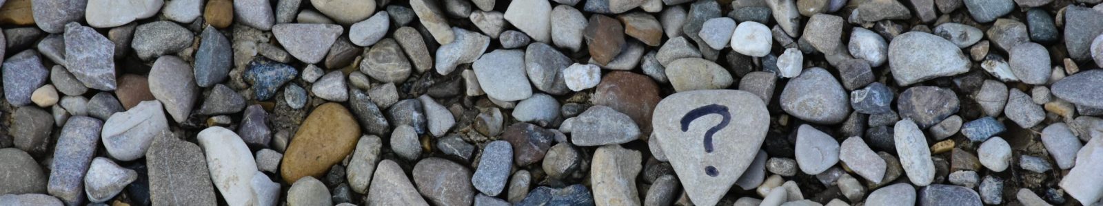 A close-up of a smooth, heart-shaped stone with a hand-drawn question mark on it, surrounded by many other pebbles.